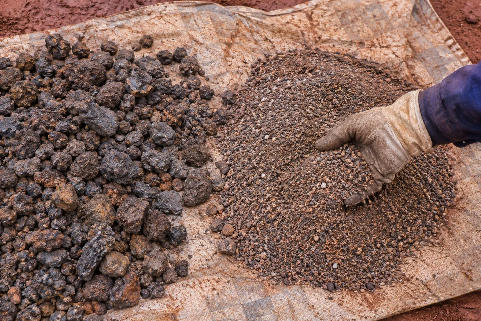 Illustrative visualisation: two separated heaps of material on a woven polypropylene sheet, coarse dark lumps on the left and screened fine granules on the right, a gloved hand drawing through the fines
