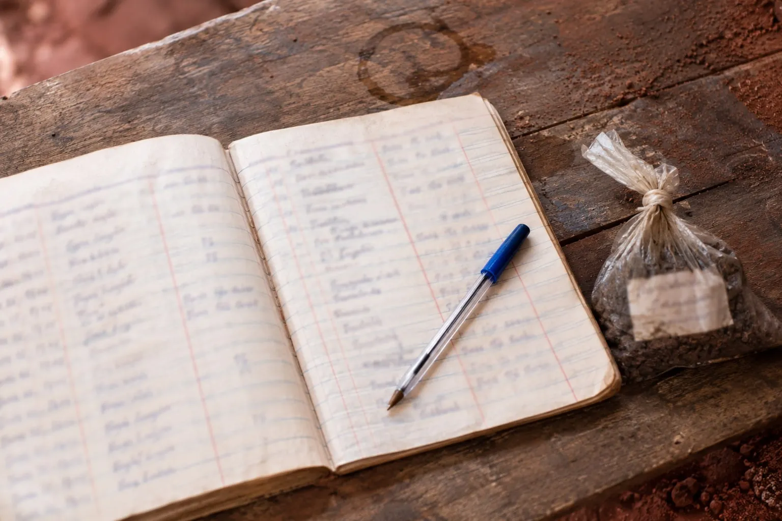 Illustrative visualisation: a handwritten paper ledger open on a weathered timber bench beside a small tied sample bag of material with a written label