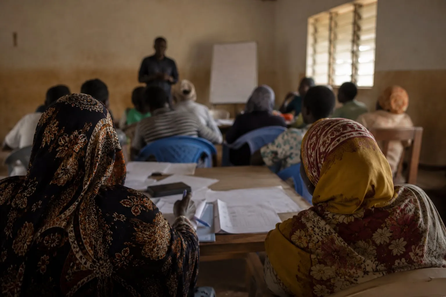 Illustrative visualisation: a training session in a plain rendered classroom, people seated around a table with printed forms and notebooks while a facilitator stands at a flip chart